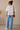 Woman wearing a white lace top and blue jeans against a beige wall.