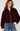 A woman wearing a cropped, deep burgundy bouclé shearling jacket with a gold zipper, paired with blue jeans and a cream ribbed sweater, against a light gray studio background.