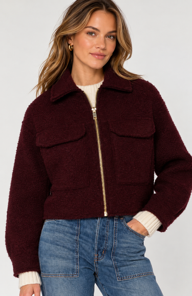 A woman wearing a cropped, deep burgundy bouclé shearling jacket with a gold zipper, paired with blue jeans and a cream ribbed sweater, against a light gray studio background.