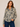 A woman modeling a long-sleeve button-up blouse with an abstract leopard-like print in beige, black, and olive green, paired with dark blue jeans against a plain beige studio background.