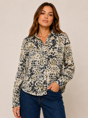 A woman modeling a long-sleeve button-up blouse with an abstract leopard-like print in beige, black, and olive green, paired with dark blue jeans against a plain beige studio background.