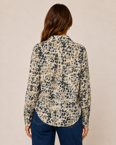 A woman viewed from the back wearing a long-sleeve button-up shirt with a beige, black, and brown floral pattern, paired with blue jeans, against a solid beige background.