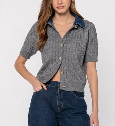 A woman modeling a short-sleeve gray cable-knit cropped cardigan with a contrasting dark blue collar and gold buttons, paired with dark blue jeans against a light gray studio background.