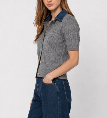A woman modeling a short-sleeve gray knit button-up shirt with a contrasting dark blue collar, paired with dark blue jeans, against a light gray studio background.