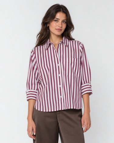 A woman modeling a long-sleeve button-up shirt with vertical burgundy and white stripes, paired with brown trousers, against a light gray studio background.