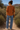 Woman in a rust-colored blouse and blue jeans standing in a desert landscape with mountains in the background.