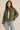 A woman wearing an olive green quilted jacket over a white ribbed top and light blue jeans, standing against a plain beige studio background.