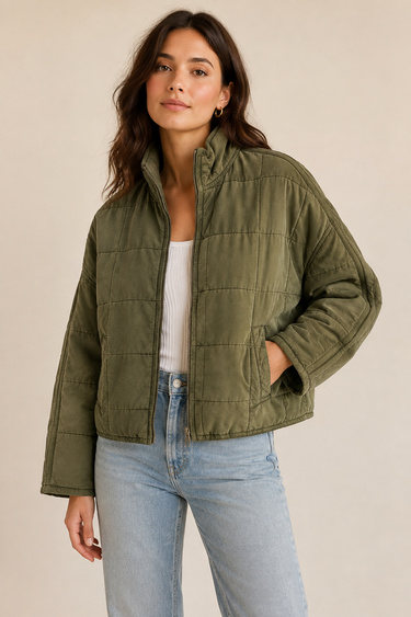 A woman wearing an olive green quilted jacket over a white ribbed top and light blue jeans, standing against a plain beige studio background.