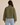 A woman viewed from the back wearing an olive green quilted jacket and light blue jeans, set against a plain beige studio background.