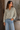 A woman wearing a long-sleeve button-up shirt with vertical olive green and cream stripes, paired with light-wash blue jeans, standing in front of a rustic wooden door and a potted plant.