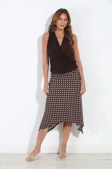 A woman modeling a sleeveless dark brown wrap-style top paired with a matching dark brown and white polka dot midi skirt, standing against a plain white wall.