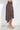 A woman wearing a brown and white polka dot high-waisted wrap skirt with an asymmetrical hem, paired with clear heeled mule sandals, standing against a white wall.