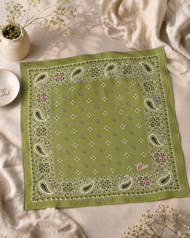An olive green bandana with a white paisley border and small white floral motifs, styled flat on a beige fabric background with dried flowers and a small dish.
