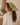 A woman wearing a sage green paisley bandana headscarf tied under her chin, paired with a white long-sleeve shirt, shown in profile against a warm-toned indoor background.