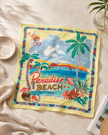 A square beach-themed scarf laid flat on a light beige fabric surface. The scarf features a colorful tropical illustration with palm trees, a blue ocean, mountains, and the words 'Paradise BEACH' in red and gold lettering.