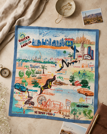 A square fabric map-style print bandanafeaturing illustrated landmarks and routes across the United States, laid flat on a beige surface with a cream blanket, small photo card, and other props around it.