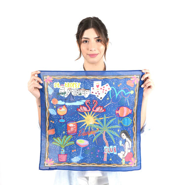 A woman holding up a square blue scarf with a colorful retro-themed graphic print featuring flamingos, drinks, cards, palm trees, and the text 'CLUB' and 'St.Tropez’