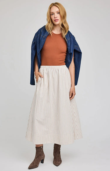 Woman wearing a rust top, white pleated skirt, and blue jacket on a plain background