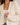 A close-up of a woman wearing a chunky cream open-front cardigan over a light beige tank top and blue jeans. The cardigan features a textured, open-knit pattern and ribbed cuffs and hem.