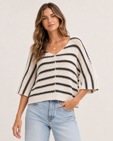 A woman wearing a cream and black horizontally striped knit sweater with elbow-length sleeves and a V-neck, paired with light blue jeans, against a plain light beige background.