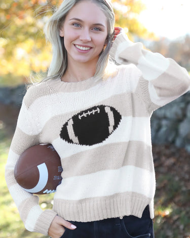 A smiling blonde woman outdoors wearing a cream and beige horizontally striped knit sweater with a large black football graphic on the chest, holding a brown football.