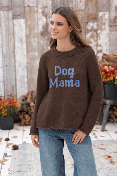 Woman wearing a brown knit “Dog Mama” sweater with light blue lettering and relaxed blue jeans in a rustic outdoor setting.