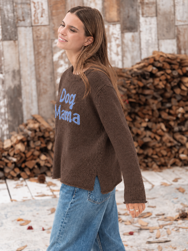 Woman wearing a brown knit “Dog Mama” sweater with light blue lettering and relaxed blue jeans in a rustic outdoor setting.