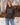 Woman wearing a brown knit “Dog Mama” sweater with light blue lettering and relaxed blue jeans in a rustic outdoor setting.