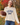 A woman holding a football wearing a cream-colored, chunky-knit long-sleeve sweater with “GAME DAY” embroidered on the front and blue jeans, photographed from behind in an outdoor autumn park with colorful fall foliage.