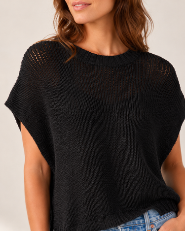 A close-up of a woman wearing a black open-knit short-sleeve top with a crew neck, paired with blue jeans, against a plain light beige background.