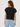 A woman shown from the back wearing a black ribbed short-sleeve knit top and light blue jeans against a plain light gray studio background.