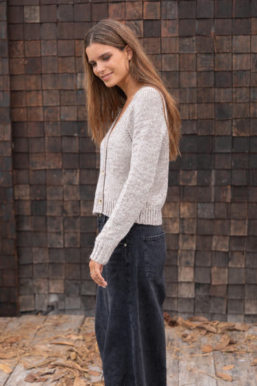 Woman wearing a light oatmeal marled V-neck button-front cardigan with black wide-leg jeans against a rustic wood shingle wall.