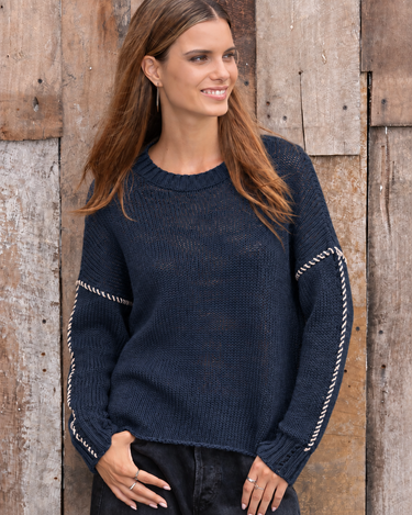 A woman wearing a navy blue knit sweater with silver rope-style trim on the sleeves, standing against a rustic wooden plank wall.
