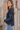 A woman wearing a navy blue ribbed knit sweater with silver contrast stitching on the sleeves, paired with light blue jeans, standing in front of a rustic wooden plank wall.