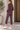 A woman modeling a matching burgundy athletic tracksuit, featuring a zip-up jacket and cropped pants, paired with white sneakers, against a outdoor setting.