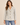 A woman modeling a light beige linen-blend blazer with brown buttons, worn over a white top and blue jeans, against a solid beige background.