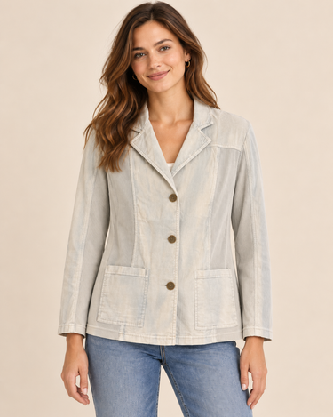 A woman modeling a light beige linen-blend blazer with brown buttons, worn over a white top and blue jeans, against a solid beige background.