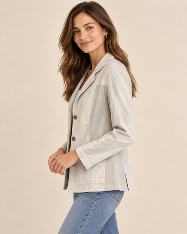 A woman modeling a light beige linen-blend blazer with brown buttons, worn over a white top and blue jeans, against a solid beige background.