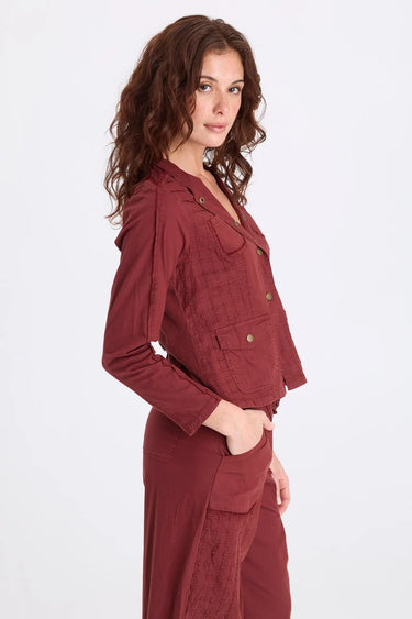 A woman facing the side wearing a blazer sports grid pintucking, rib contrast panels and brass hardware and matching berry pants on a white background.