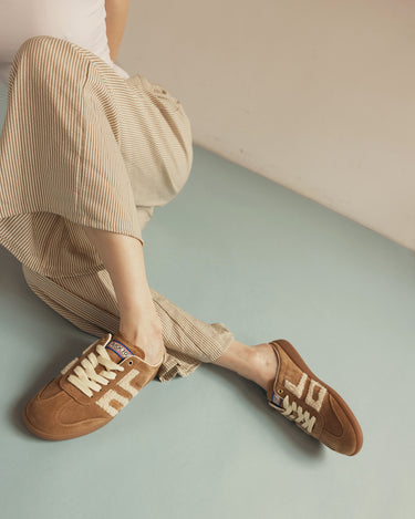 A person wearing beige striped pants and brown suede sneakers with cream lettering, seated against a pale blue floor and cream wall.
