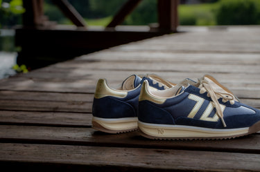 A pair of navy blue and cream New Balance 700 sneakers resting on a weathered wooden deck with a blurred outdoor background.