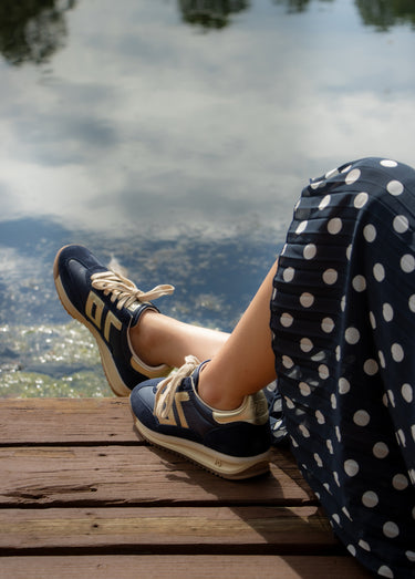 A person sitting on a wooden dock by the water, wearing navy blue sneakers with beige stripes and a navy skirt with white polka dots.