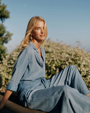 A woman wearing a light blue denim shirt and jeanssits outdoors on a wooden bench with green bushes and a clear blue sky in the background.