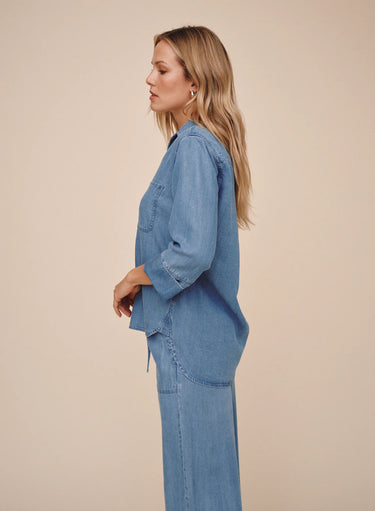 A woman in side profile wearing a light blue denim long-sleeve shirt and matching wide-leg pants against a solid beige studio background.