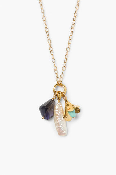 Gold chain necklace with a cluster of charms, including a long baroque pearl, a faceted dark purple iolite gemstone, and a gold floral charm with a small turquoise bead, presented on a white background.