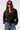A woman modeling a black belted double-breasted jacket with dark sunglasses and blue jeans, standing against a plain light background.