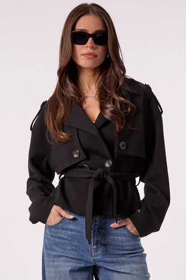 A woman modeling a black belted double-breasted jacket with dark sunglasses and blue jeans, standing against a plain light background.