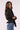 A woman wearing a black long-sleeve wrap-style blouse with a tied waist and blue jeans, photographed against a plain light background.