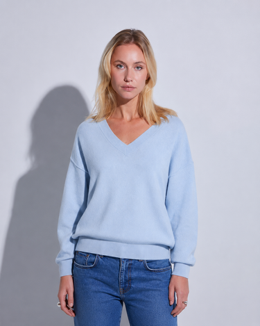 A blonde woman wearing a light blue v-neck sweater and blue jeans, standing against a light gray wall with a soft shadow cast to the side.