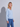 A blonde woman wearing a light blue v-neck knit sweater and blue jeans, standing against a light gray studio background with a soft shadow cast to the left.